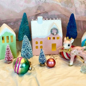 K&ouml;nnte beinhalten: Eine festliche Weihnachtsdekoration mit Miniaturh&auml;usern in Gr&uuml;n, Rosa und Blau mit schneebedeckten D&auml;chern. Die Szene umfasst Flaschenb&uuml;rstenb&auml;ume, Ornamente und eine rosa Hirschfigur, die ein charmantes Weihnachtsdorf bilden.