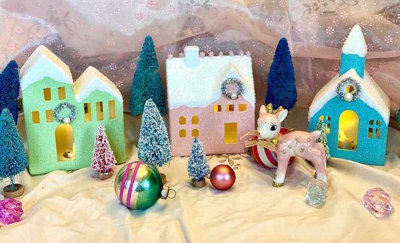 K&ouml;nnte beinhalten: Eine festliche Weihnachtsdekoration mit Miniaturh&auml;usern in Gr&uuml;n, Rosa und Blau mit schneebedeckten D&auml;chern. Die Szene umfasst Flaschenb&uuml;rstenb&auml;ume, Ornamente und eine rosa Hirschfigur, die ein charmantes Weihnachtsdorf bilden.