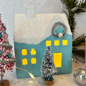K&ouml;nnte beinhalten: Eine festliche Weihnachtsdorf-Szene mit einem hellblauen glitzernden Haus, einem schneebedeckten Dach und einem silbernen Kranz. Rote und blaue Flaschenb&uuml;rstenb&auml;ume, verziert mit Ornamenten, umgeben das Haus. Eine Lichterkette und silberne Kugeln vervollst&auml;ndigen die festliche Pr&auml;sentation.