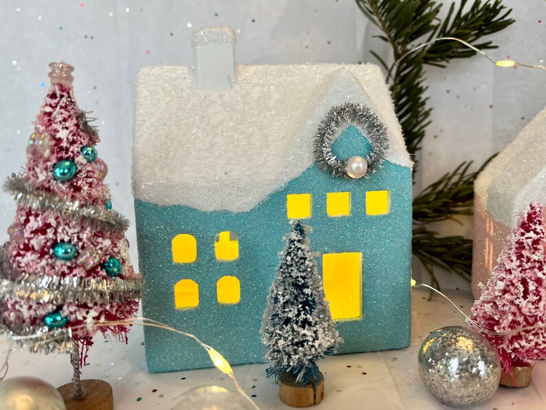 K&ouml;nnte beinhalten: Eine festliche Weihnachtsdorf-Szene mit einem hellblauen glitzernden Haus, einem schneebedeckten Dach und einem silbernen Kranz. Rote und blaue Flaschenb&uuml;rstenb&auml;ume, verziert mit Ornamenten, umgeben das Haus. Eine Lichterkette und silberne Kugeln vervollst&auml;ndigen die festliche Pr&auml;sentation.