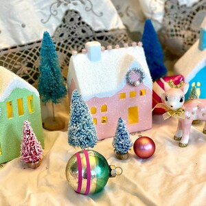 K&ouml;nnte beinhalten: Eine festliche Urlaubsszene mit Miniaturh&auml;usern in Pastellfarben, Flaschenb&uuml;rstenb&auml;umen und einem rosa Keramikhirsch mit goldener Krone. Dekorative Ornamente und eine Vintage-&Auml;sthetik schaffen eine bezaubernde Weihnachtsausstellung.