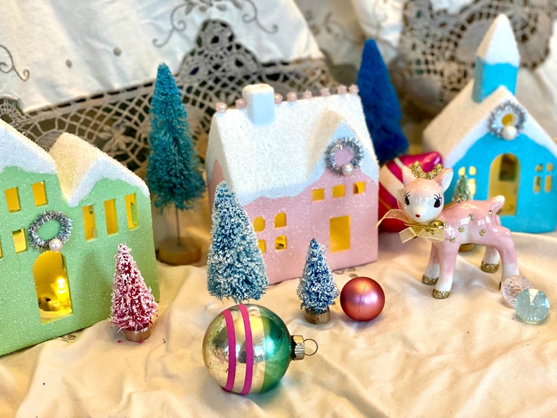 K&ouml;nnte beinhalten: Eine festliche Urlaubsszene mit Miniaturh&auml;usern in Pastellfarben, Flaschenb&uuml;rstenb&auml;umen und einem rosa Keramikhirsch mit goldener Krone. Dekorative Ornamente und eine Vintage-&Auml;sthetik schaffen eine bezaubernde Weihnachtsausstellung.