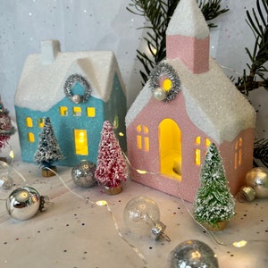 K&ouml;nnte beinhalten: Eine festliche Urlaubsszene mit zwei beleuchteten Keramikh&auml;usern, einem blauen und einem rosa, mit glitzernden, schneebedeckten D&auml;chern. Miniatur-Flaschenb&uuml;rstenb&auml;ume und silberne Ornamente umgeben die H&auml;user und schaffen ein charmantes Winterdorf.