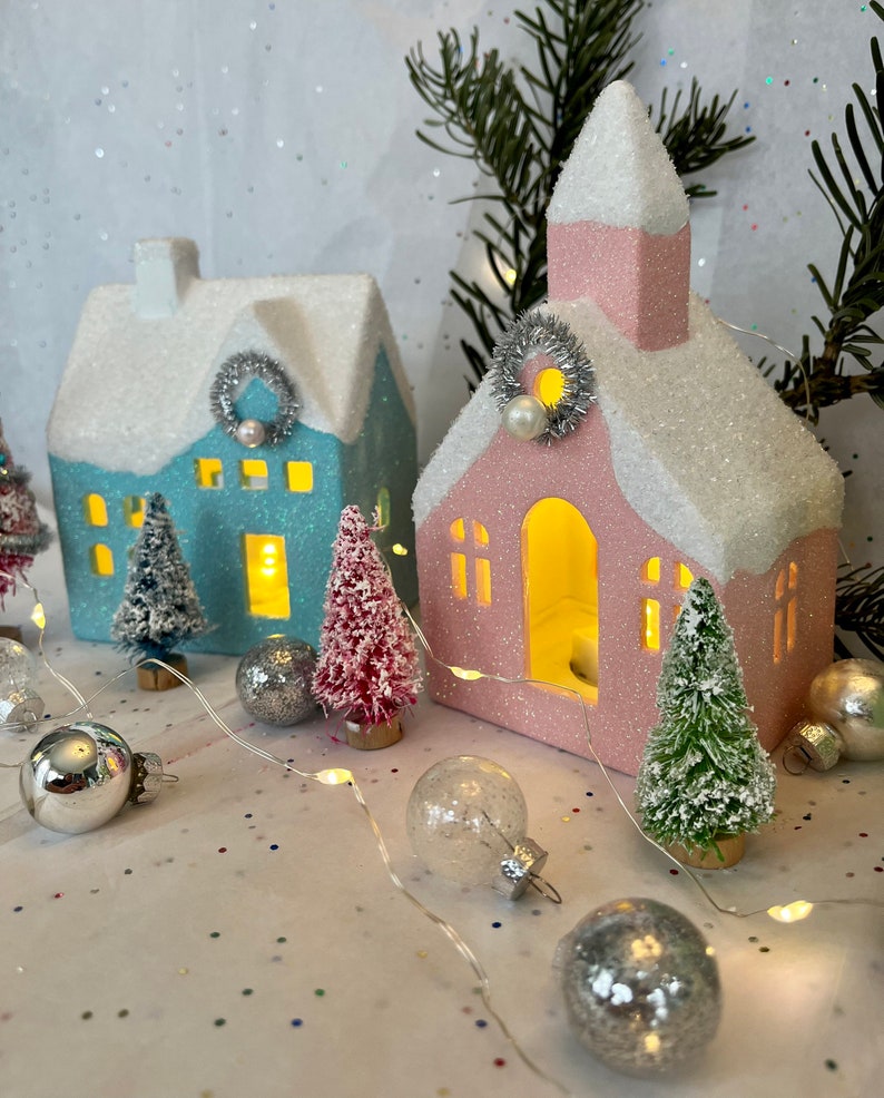 K&ouml;nnte beinhalten: Eine festliche Urlaubsszene mit zwei beleuchteten Keramikh&auml;usern, einem blauen und einem rosa, mit glitzernden, schneebedeckten D&auml;chern. Miniatur-Flaschenb&uuml;rstenb&auml;ume und silberne Ornamente umgeben die H&auml;user und schaffen ein charmantes Winterdorf.