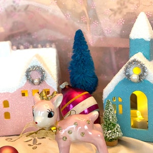 K&ouml;nnte beinhalten: Eine festliche Weihnachtsdekoration mit einem rosa Haus und einer blauen Kirche, beide mit schneebedeckten D&auml;chern, silbernen Kr&auml;nzen und Perlenakzenten. Eine rosa Hirschfigur mit goldener Krone und ein Vintage-Ornament tragen zum festlichen Charme bei. Miniatur blaue und gr&uuml;ne B&auml;ume vervollst&auml;ndigen die Szene.