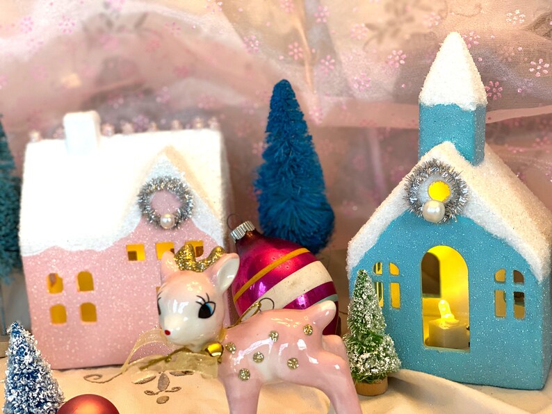 K&ouml;nnte beinhalten: Eine festliche Weihnachtsdekoration mit einem rosa Haus und einer blauen Kirche, beide mit schneebedeckten D&auml;chern, silbernen Kr&auml;nzen und Perlenakzenten. Eine rosa Hirschfigur mit goldener Krone und ein Vintage-Ornament tragen zum festlichen Charme bei. Miniatur blaue und gr&uuml;ne B&auml;ume vervollst&auml;ndigen die Szene.