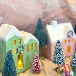 K&ouml;nnte beinhalten: Eine festliche Weihnachtsdorf-Szene mit Miniaturh&auml;usern in Pastellgr&uuml;n und -rosa, gekr&ouml;nt von schneewei&szlig;en D&auml;chern. Die H&auml;user sind mit Kr&auml;nzen geschm&uuml;ckt und von kleinen, beflockten Weihnachtsb&auml;umen und Kugeln umgeben. Ein warmes Licht scheint aus dem Inneren der H&auml;user.