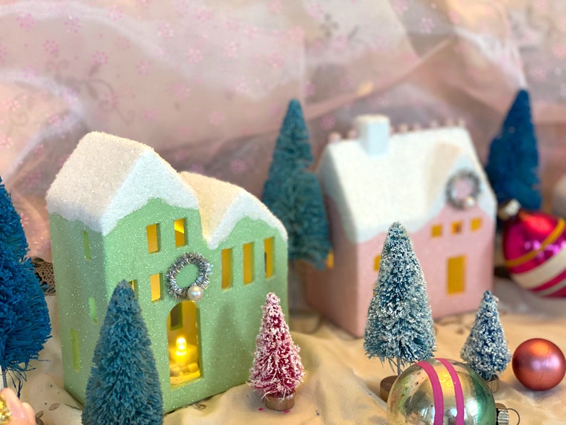 K&ouml;nnte beinhalten: Eine festliche Weihnachtsdorf-Szene mit Miniaturh&auml;usern in Pastellgr&uuml;n und -rosa, gekr&ouml;nt von schneewei&szlig;en D&auml;chern. Die H&auml;user sind mit Kr&auml;nzen geschm&uuml;ckt und von kleinen, beflockten Weihnachtsb&auml;umen und Kugeln umgeben. Ein warmes Licht scheint aus dem Inneren der H&auml;user.