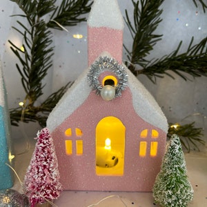 K&ouml;nnte beinhalten: Eine rosa kirchenf&ouml;rmige Weihnachtsdekoration mit schneebedecktem Dach und silbernem Kranz. Die Kirche hat einen beleuchteten Innenraum und wird von kleinen Weihnachtsb&auml;umen und Ornamenten flankiert. Eine Lichterkette und Tannenzweige tragen zur festlichen Szene bei.