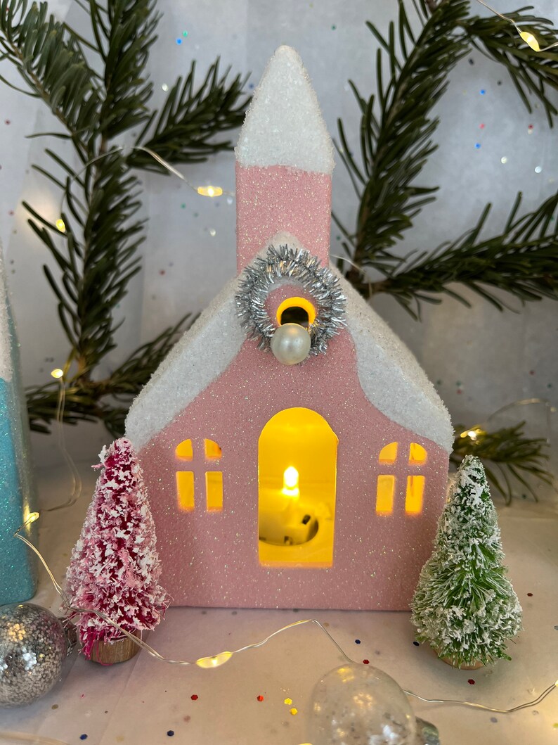 K&ouml;nnte beinhalten: Eine rosa kirchenf&ouml;rmige Weihnachtsdekoration mit schneebedecktem Dach und silbernem Kranz. Die Kirche hat einen beleuchteten Innenraum und wird von kleinen Weihnachtsb&auml;umen und Ornamenten flankiert. Eine Lichterkette und Tannenzweige tragen zur festlichen Szene bei.