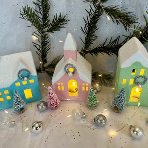 K&ouml;nnte beinhalten: Eine festliche Weihnachtsszene mit drei beleuchteten Keramikh&auml;usern in Blau, Rosa und Gr&uuml;n, mit schneebedeckten D&auml;chern. Miniatur-Weihnachtsb&auml;ume und silberne Ornamente umgeben die H&auml;user, mit einer Lichterkette. Eine charmante Winterdorf-Dekoration.