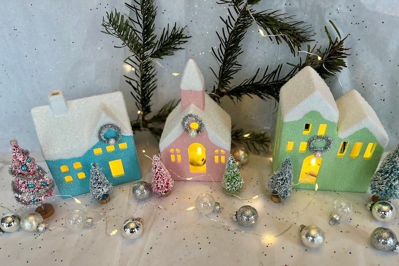 K&ouml;nnte beinhalten: Eine festliche Weihnachtsszene mit drei beleuchteten Keramikh&auml;usern in Blau, Rosa und Gr&uuml;n, mit schneebedeckten D&auml;chern. Miniatur-Weihnachtsb&auml;ume und silberne Ornamente umgeben die H&auml;user, mit einer Lichterkette. Eine charmante Winterdorf-Dekoration.