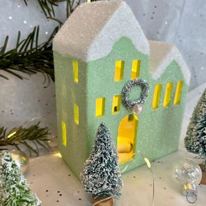 K&ouml;nnte beinhalten: Ein hellgr&uuml;nes, glitzerbedecktes dekoratives Haus mit schneewei&szlig;em Dach und gelb leuchtenden Fenstern. Ein kleiner silberner Kranz und eine Perle schm&uuml;cken die Vorderseite. Miniatur-Weihnachtsb&auml;ume und Ornamente umgeben das Haus, mit einer Lichterkette.