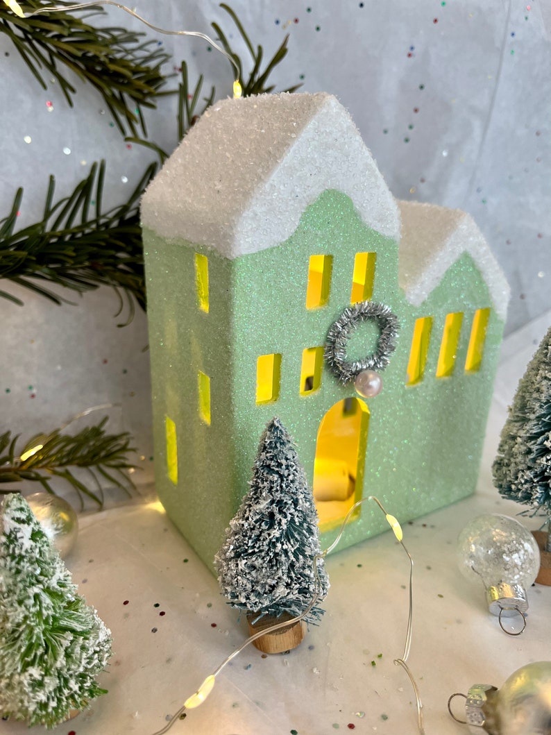 K&ouml;nnte beinhalten: Ein hellgr&uuml;nes, glitzerbedecktes dekoratives Haus mit schneewei&szlig;em Dach und gelb leuchtenden Fenstern. Ein kleiner silberner Kranz und eine Perle schm&uuml;cken die Vorderseite. Miniatur-Weihnachtsb&auml;ume und Ornamente umgeben das Haus, mit einer Lichterkette.