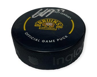 May include: Black official game puck with the text "Bruins 100 Centennial 1924-2024" in gold and white. The puck has a textured edge and a signature in white. The National Hockey League logo is visible.