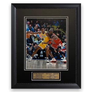 May include: Framed photograph featuring Kobe Bryant and Michael Jordan in action during a basketball game. Bryant is wearing a yellow Lakers jersey, while Jordan is in a red Bulls uniform. The frame is black with a decorative border, and includes nameplates.