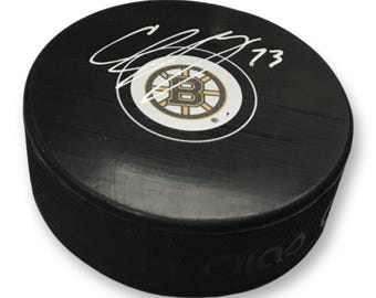 May include: Black hockey puck featuring the Boston Bruins logo in a white circle with gold and black accents. The puck has a signature and the number 73 in white. The puck is a standard size for ice hockey.