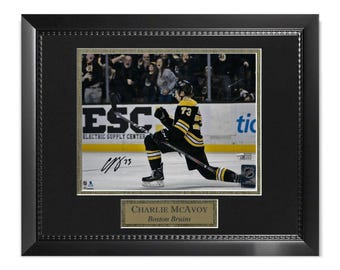 May include: Framed photo of hockey player Charlie McAvoy of the Boston Bruins. McAvoy is kneeling on the ice in his black and gold uniform, celebrating a goal. The frame is black with a gold border and a nameplate.
