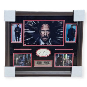 May include: Framed John Wick movie memorabilia featuring multiple images of Keanu Reeves, including a central portrait surrounded by guns. The frame includes a signature and a plaque that reads "JOHN WICK KEANU REEVES."