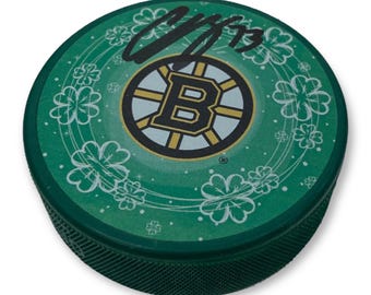 May include: A green hockey puck featuring the Boston Bruins logo in the center, surrounded by white shamrocks. The puck has a dark green edge and a signature with the number 13. The team logo is black, gold, and white.