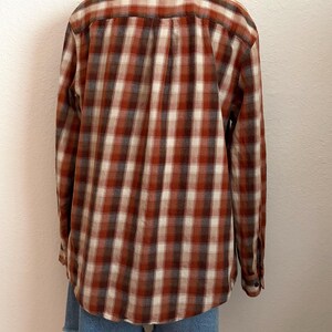 May include: A long-sleeve button-down shirt featuring a plaid pattern in shades of rust orange, cream, and dark brown. The shirt has a standard collar, a back yoke with a center pleat, and buttoned cuffs. It is paired with blue denim shorts.