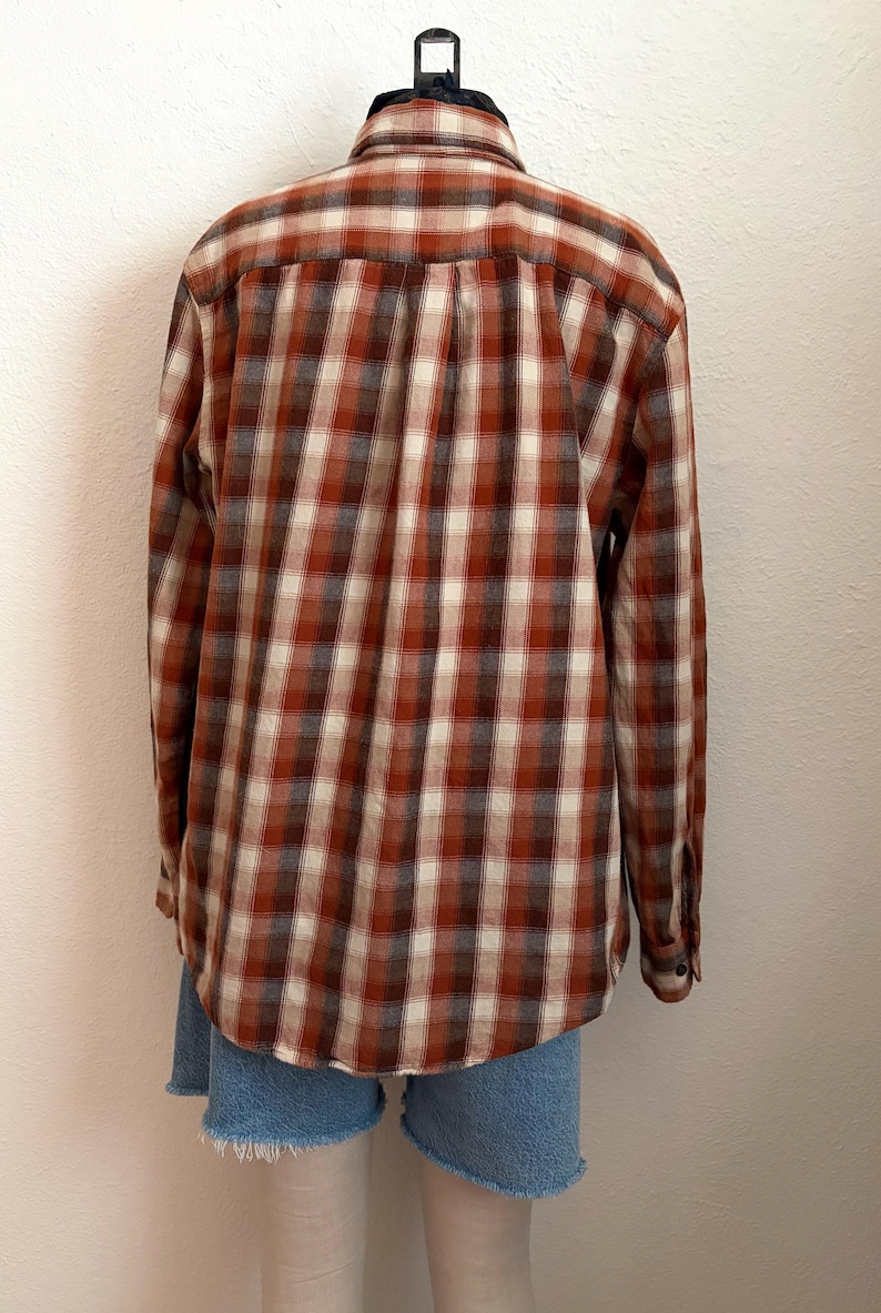 May include: A long-sleeve button-down shirt featuring a plaid pattern in shades of rust orange, cream, and dark brown. The shirt has a standard collar, a back yoke with a center pleat, and buttoned cuffs. It is paired with blue denim shorts.