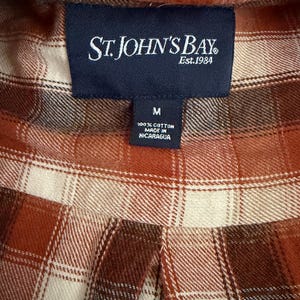 May include: A long-sleeve shirt featuring a plaid pattern in shades of rust orange, cream, and dark brown. A dark blue rectangular label at the collar reads St. John's Bay Est. 1984, with a smaller tag below indicating size M, 100% cotton, and made in Nicaragua.