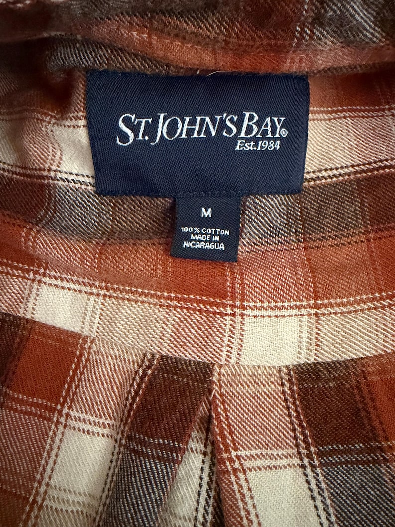 May include: A long-sleeve shirt featuring a plaid pattern in shades of rust orange, cream, and dark brown. A dark blue rectangular label at the collar reads St. John's Bay Est. 1984, with a smaller tag below indicating size M, 100% cotton, and made in Nicaragua.