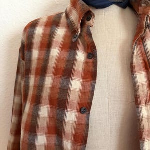 May include: A long-sleeve button-down shirt featuring a plaid pattern in shades of burnt orange, cream, and dark brown. The shirt has a pointed collar and dark buttons down the front placket. A dark blue paisley patterned neckerchief is tied at the neck.