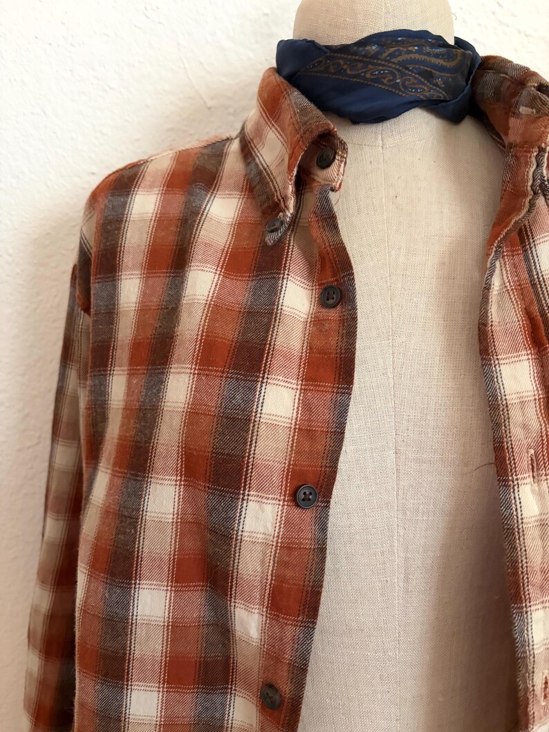 May include: A long-sleeve button-down shirt featuring a plaid pattern in shades of burnt orange, cream, and dark brown. The shirt has a pointed collar and dark buttons down the front placket. A dark blue paisley patterned neckerchief is tied at the neck.