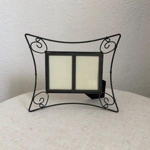 May include: A double photo frame featuring a rectangular central display with two vertical slots, encased in a dark metal wire structure. The frame has curved sides and decorative scrollwork accents at each of the four corners of the wire border.