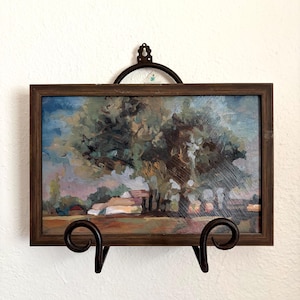 May include: A rectangular oil painting in a dark brown wooden frame, depicting a landscape with large green trees, a small house, and a field under a blue sky. The artwork is displayed on a black metal scrollwork stand with a wall-mounted hook.