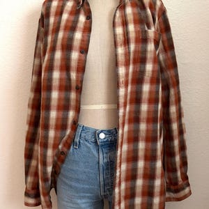 May include: A long-sleeve button-down shirt featuring a plaid pattern in shades of rust orange, cream, and dark brown. The shirt is worn over light blue denim shorts with a raw hem. A dark blue neckerchief is tied around the neck of the mannequin.