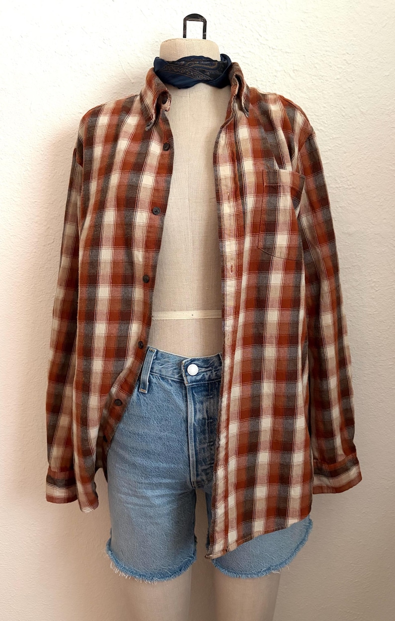 May include: A long-sleeve button-down shirt featuring a plaid pattern in shades of rust orange, cream, and dark brown. The shirt is worn over light blue denim shorts with a raw hem. A dark blue neckerchief is tied around the neck of the mannequin.