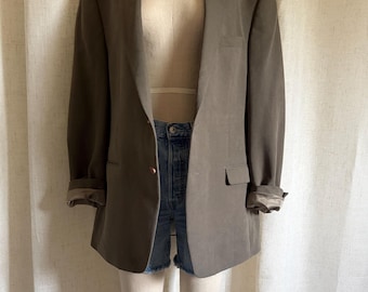 May include: A taupe blazer with notched lapels, two front buttons, and two flap pockets, worn over blue denim shorts. The blazer features long sleeves with rolled cuffs, revealing a smooth, light-colored lining, and a dark neck accessory.