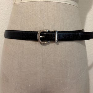 Puede incluir: Un cinturón negro con superficie texturizada y hebilla de metal plateado. El cinturón cuenta con un cierre de una sola púa y una trabilla de metal plateado a juego, asegurado alrededor de un torso de maniquí de color neutro.