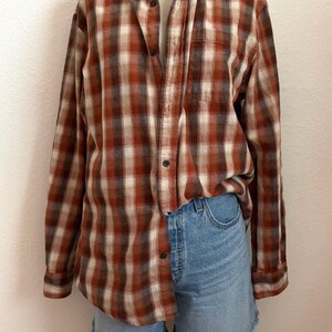 May include: A long-sleeve button-down shirt featuring a plaid pattern in shades of rust orange, cream, and dark brown, paired with light blue denim shorts that have a frayed hemline. The shirt includes a single chest pocket and dark buttons.
