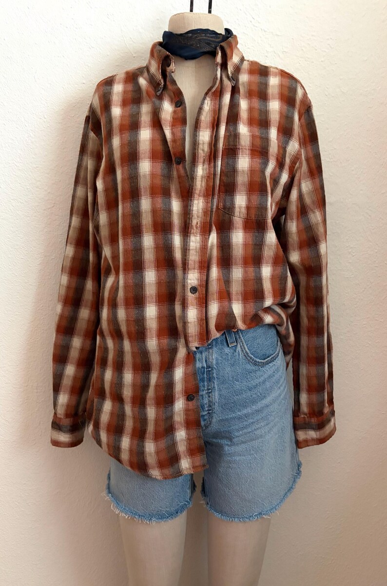 May include: A long-sleeve button-down shirt featuring a plaid pattern in shades of rust orange, cream, and dark brown, paired with light blue denim shorts that have a frayed hemline. The shirt includes a single chest pocket and dark buttons.