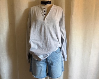 May include: A light gray long-sleeve henley shirt with a three-button placket and a black neck ribbon, paired with light blue denim shorts featuring a raw, frayed hemline. The outfit is displayed on a mannequin against a neutral fabric backdrop.