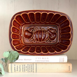 May include: A rectangular, dark brown glazed ceramic baking mold with a fluted rim and a central embossed design featuring a pumpkin flanked by an apple and a pear. The mold is resting on top of two stacked books with visible text on their spines.