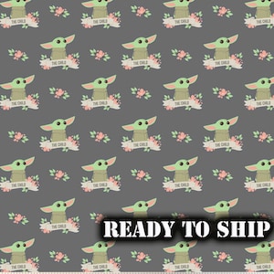 May include: A gray fabric with a repeating pattern of a green alien character with big eyes, surrounded by pink flowers and a banner that reads "The Child".