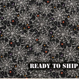 May include: Black fabric with a white spiderweb pattern, orange stars, and purple crescent moons. The text "READY TO SHIP" is displayed in a white font. The fabric is measured in inches.