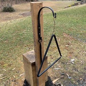 May include: A black metal triangle musical instrument hanging from a wooden post with a leather strap. The triangle is suspended by a chain. The post is mounted on a wooden block and a tree stump.