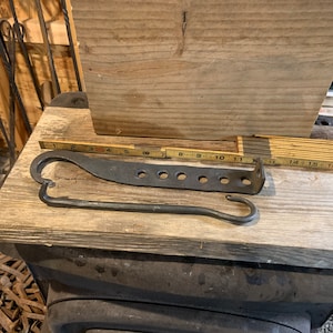 May include: Hand-forged metal tools, including a poker with a curved handle and a tool with multiple holes, rest on a wooden surface. A wooden ruler is also present. The tools appear to be for a fireplace or blacksmithing.
