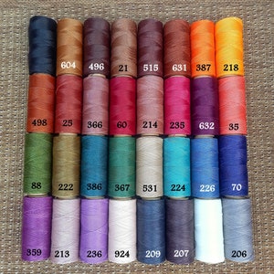 Könnte beinhalten: Eine Sammlung von 24 Spulen gewachsten Fadens in verschiedenen Farben, darunter Braun, Rot, Grün, Blau, Lila und Grau. Jede Spule ist mit einer Nummer gekennzeichnet.