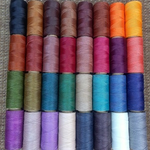Pode incluir: Uma cole&ccedil;&atilde;o de 36 carret&eacute;is de fio encerado em v&aacute;rias cores, incluindo preto, marrom, laranja, vermelho, roxo, verde, azul, cinza e branco. Os carret&eacute;is est&atilde;o dispostos em uma grade sobre uma superf&iacute;cie tecida.