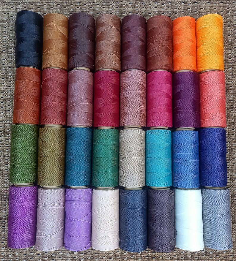 Pode incluir: Uma cole&ccedil;&atilde;o de 36 carret&eacute;is de fio encerado em v&aacute;rias cores, incluindo preto, marrom, laranja, vermelho, roxo, verde, azul, cinza e branco. Os carret&eacute;is est&atilde;o dispostos em uma grade sobre uma superf&iacute;cie tecida.