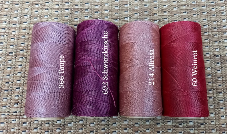 May include: Four spools of thread in different shades of pink, red, and purple. The spools are labeled with numbers and names: 366 Taupe, 692 Schwarzkirsche, 214 Altrose, and 60 Weinrot.