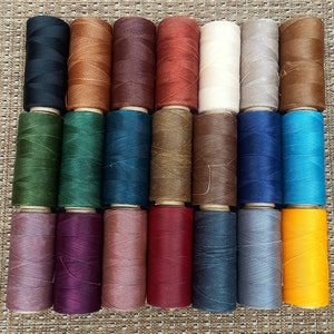 May include: A collection of 24 spools of thread in various colors, including black, brown, red, green, blue, and yellow. The spools are arranged in two rows of 12.