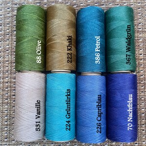 Pode incluir: Oito rolos de fio em v&aacute;rias cores, incluindo verde oliva, c&aacute;qui, azul-turquesa, azul petr&oacute;leo, bege, turquesa, azul claro e azul escuro. Cada rolo tem um n&uacute;mero e um nome de cor impresso no r&oacute;tulo.